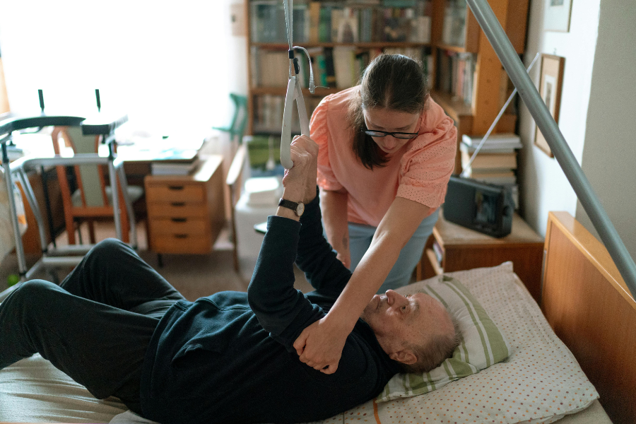 Opiekunka seniora w pracy - pomaga odwrócić się leżącemu seniorowi na łóżku.